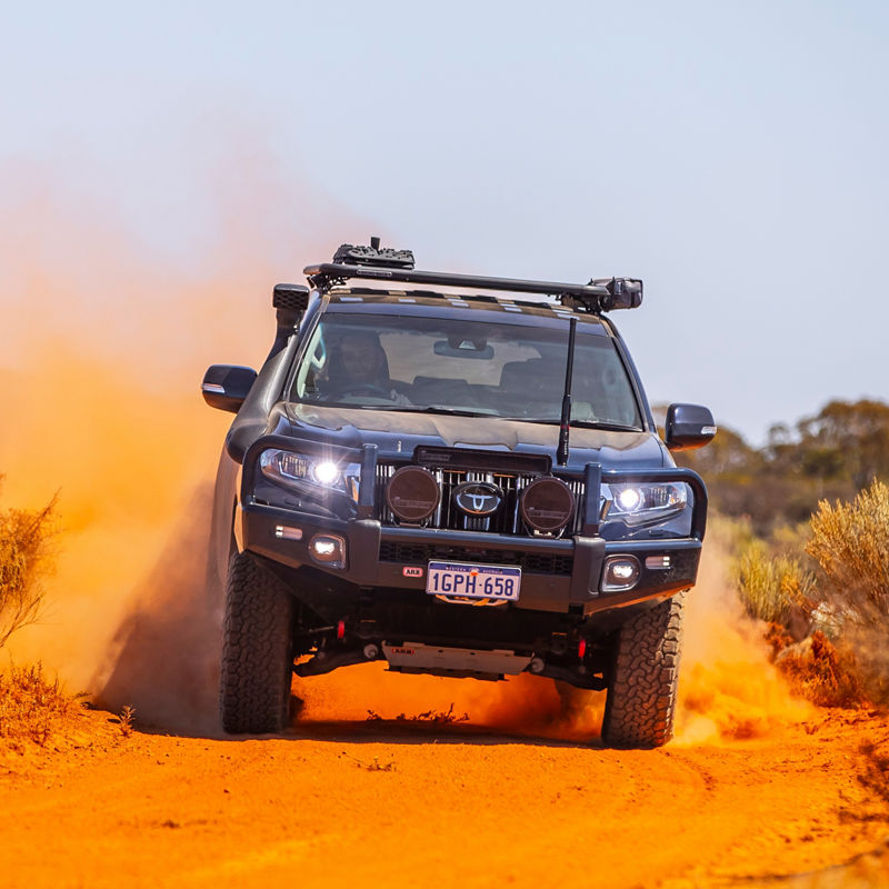Front view of a Toyota LandCruiser Prado 150 Series driving through red dust, fitted with an ARB bull bar, driving lights, roof rack system and under vehicle protection.