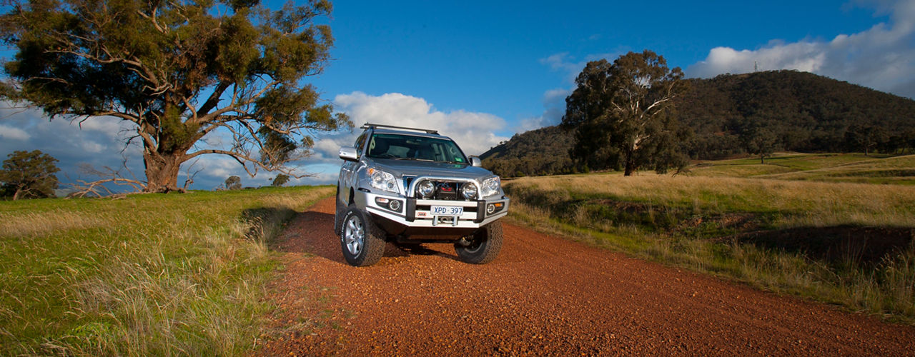 Toyota LandCruiser Prado 120 Series fitted with an ARB bull bar, driving lights, roof rack system and winch on a gravel road through rolling farmland.