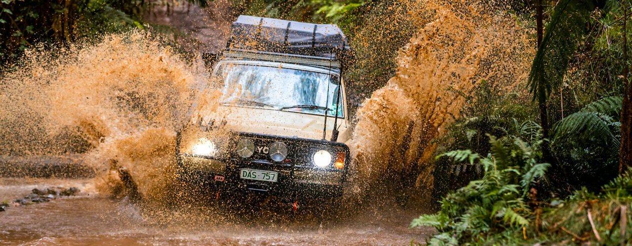 Front view of a Toyota HiLux fitted with an ARB bull bar, driving lights, snorkel and roof rack, driving through a muddy water crossing in dense forest.