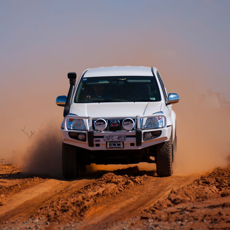 Front view of a Toyota LandCruiser Prado 90 Series driving through red dust, fitted with an ARB bull bar, driving lights and snorkel.