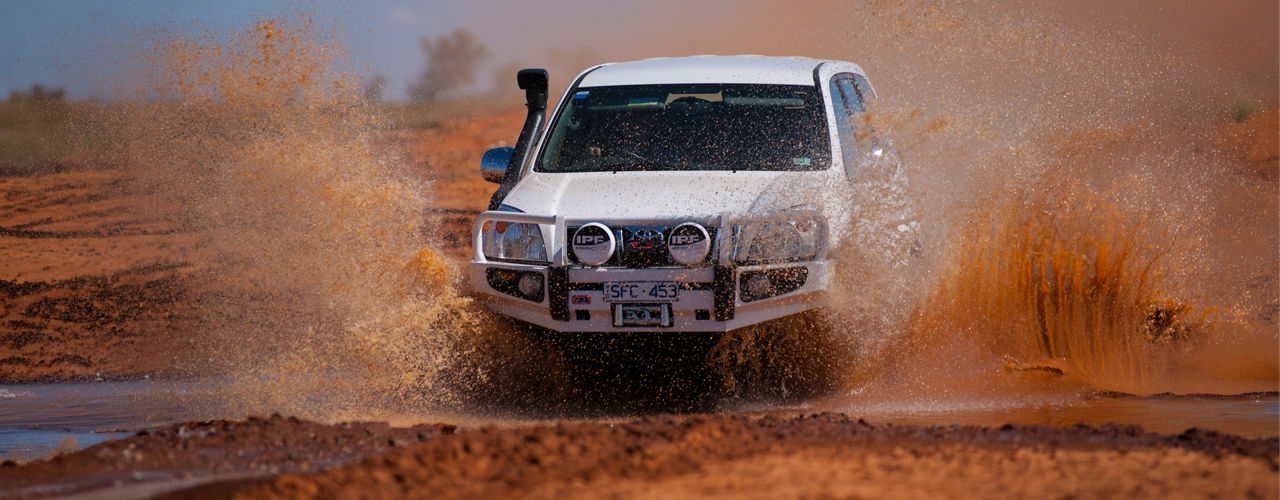 Front view of a Toyota LandCruiser Prado 90 Series driving through a muddy water crossing, fitted with an ARB bull bar, driving lights and snorkel, with water splashing around the vehicle.