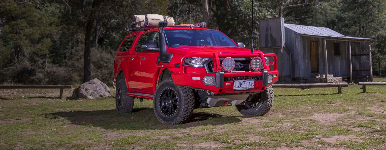 Front three-quarter view of a Ford Ranger PXII fitted with an ARB bull bar, driving lights, snorkel, roof rack and under-vehicle protection, parked beside a bush campsite.