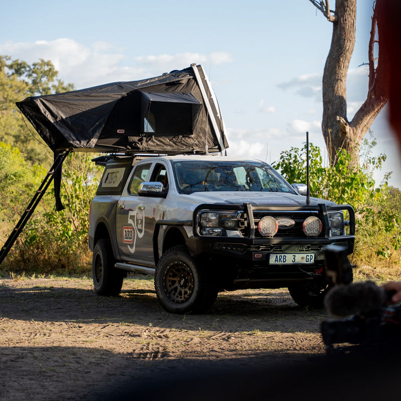 Ford Ranger with an ARB rooftop tent set up at a campsite, viewed through the frame of a camera in the foreground.