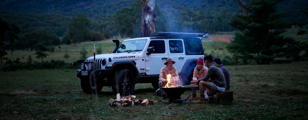 Wide view of four people gathered around a campfire beside a Jeep Wrangler Rubicon fitted with an ARB awning and driving lights in a mountain valley campsite.
