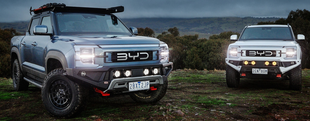 Two BYD Shark 6 vehicles fitted with ARB accessories parked on a grassy ridgeline beneath an overcast sky, with driving lights illuminated and a roof rack system visible on the foreground vehicle.