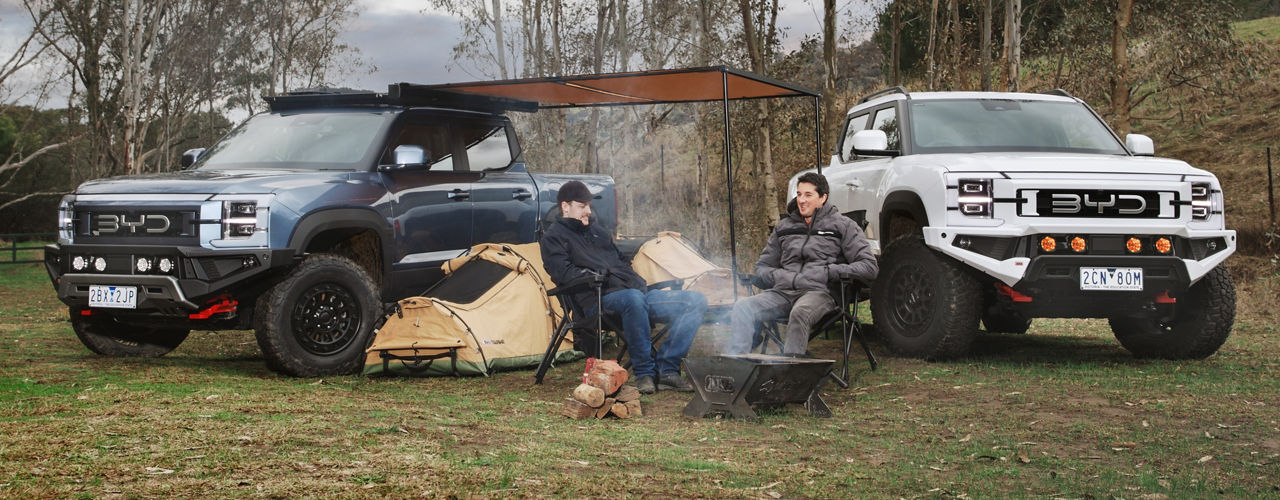 Two BYD Shark 6 vehicles fitted with ARB accessories parked at a campsite, with two people seated beside a campfire beneath an awning in a rural landscape.