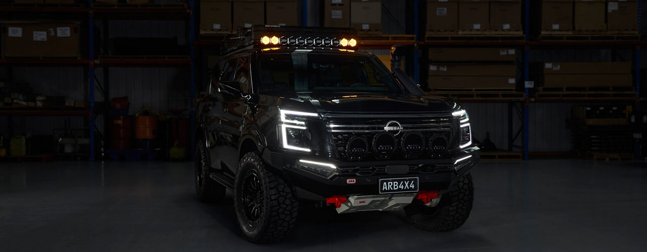 Front view of a Nissan Patrol Y63 fitted with an ARB bull bar, driving lights, roof rack system and under vehicle protection, parked inside a warehouse with lighting illuminated.