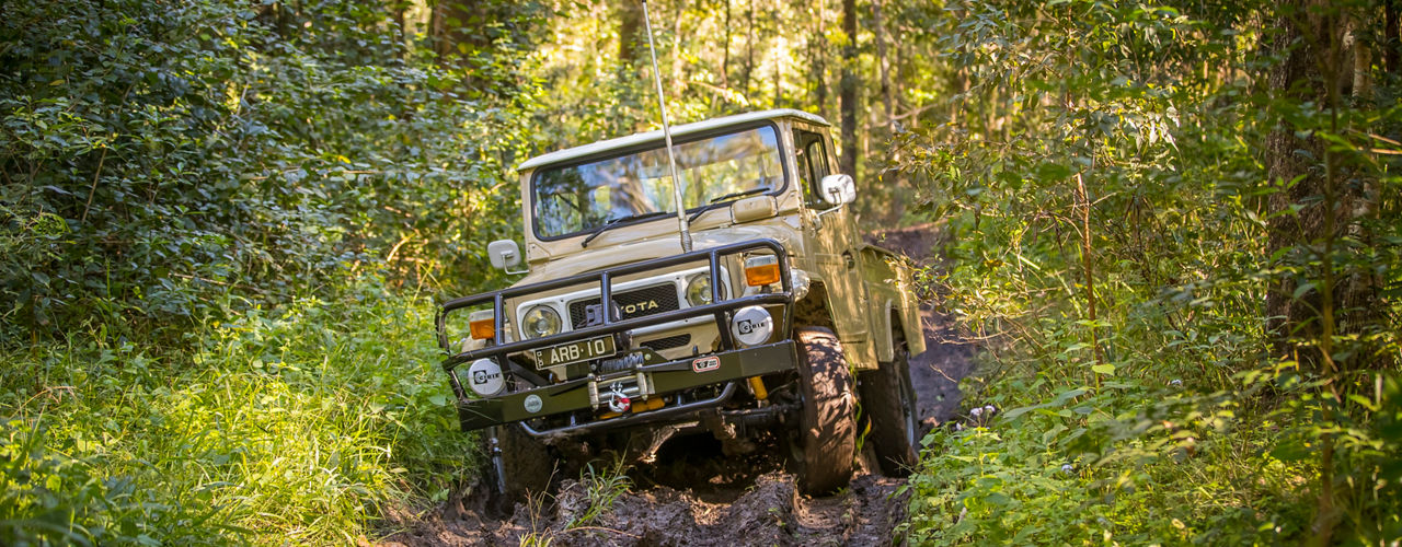Toyota LandCruiser FJ45 ute fitted with an ARB bull bar and winch, navigating a muddy forest track with visible wheel articulation.