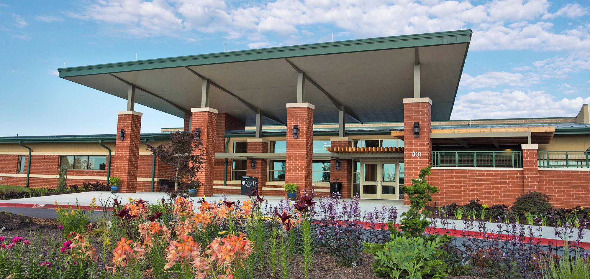 Contemporary community center facility with a broad canopy roof, brick-and-glass facade and vibrant landscaped gardens framing the main entrance beneath a bright blue sky.