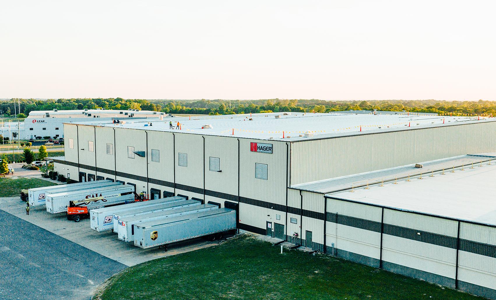 Wide aerial view of a large white industrial warehouse with a bright white roof at golden sunset, surrounded by green landscaping, loading docks and neighboring commercial buildings.
