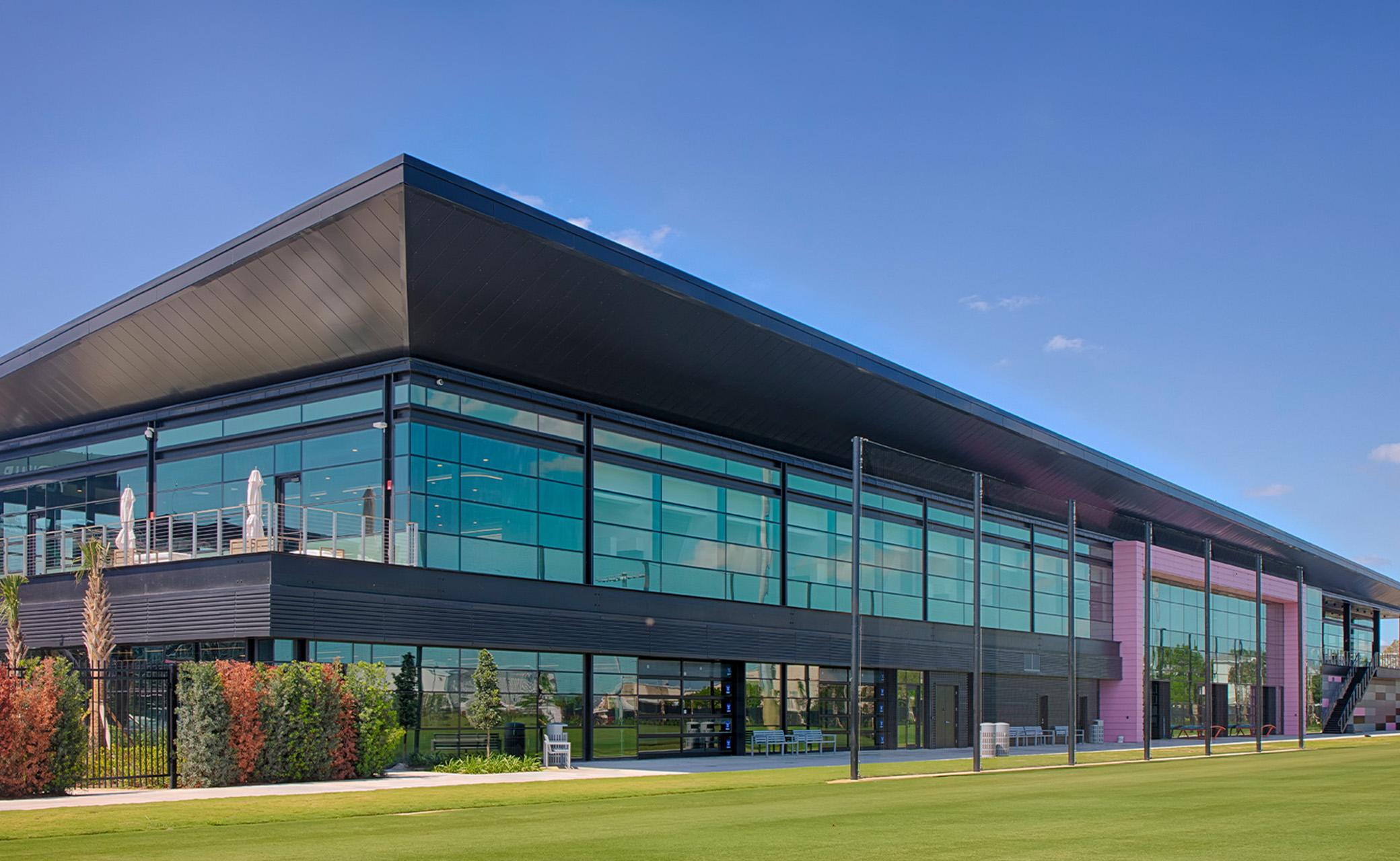 Contemporary glass-front office building with dark metal framing, vibrant accent panels and expansive green space beneath a clear blue sky.