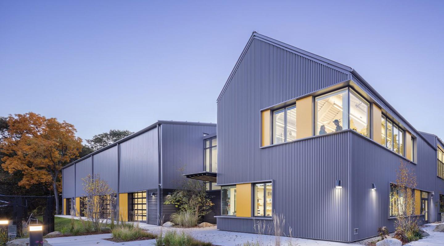 Contemporary dark gray office building with angular rooflines, expansive corner windows and warm interior lighting at dusk.