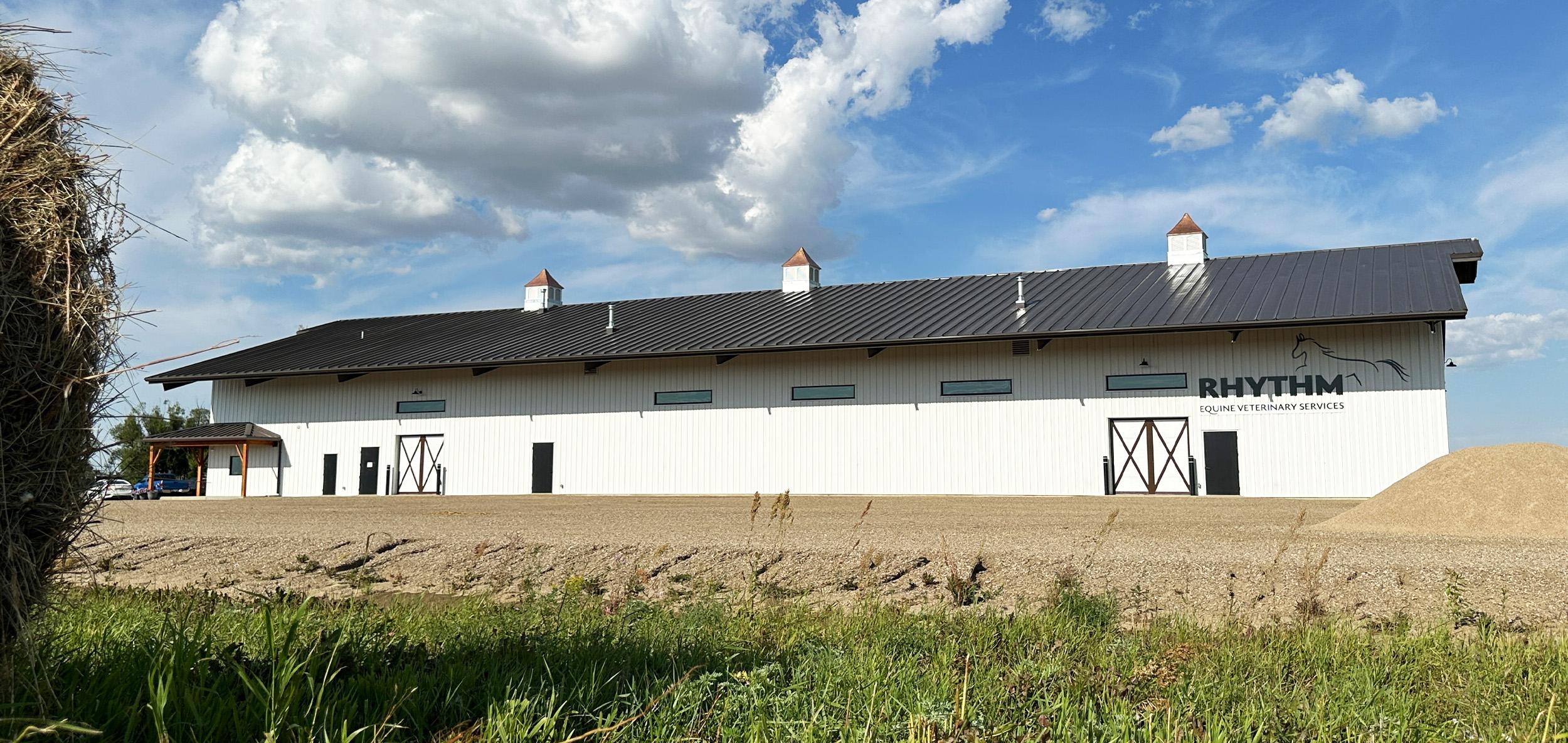 Long white agricultural facility with black roof, barn-style entry doors and open rural landscape beneath a bright blue sky with dramatic clouds.