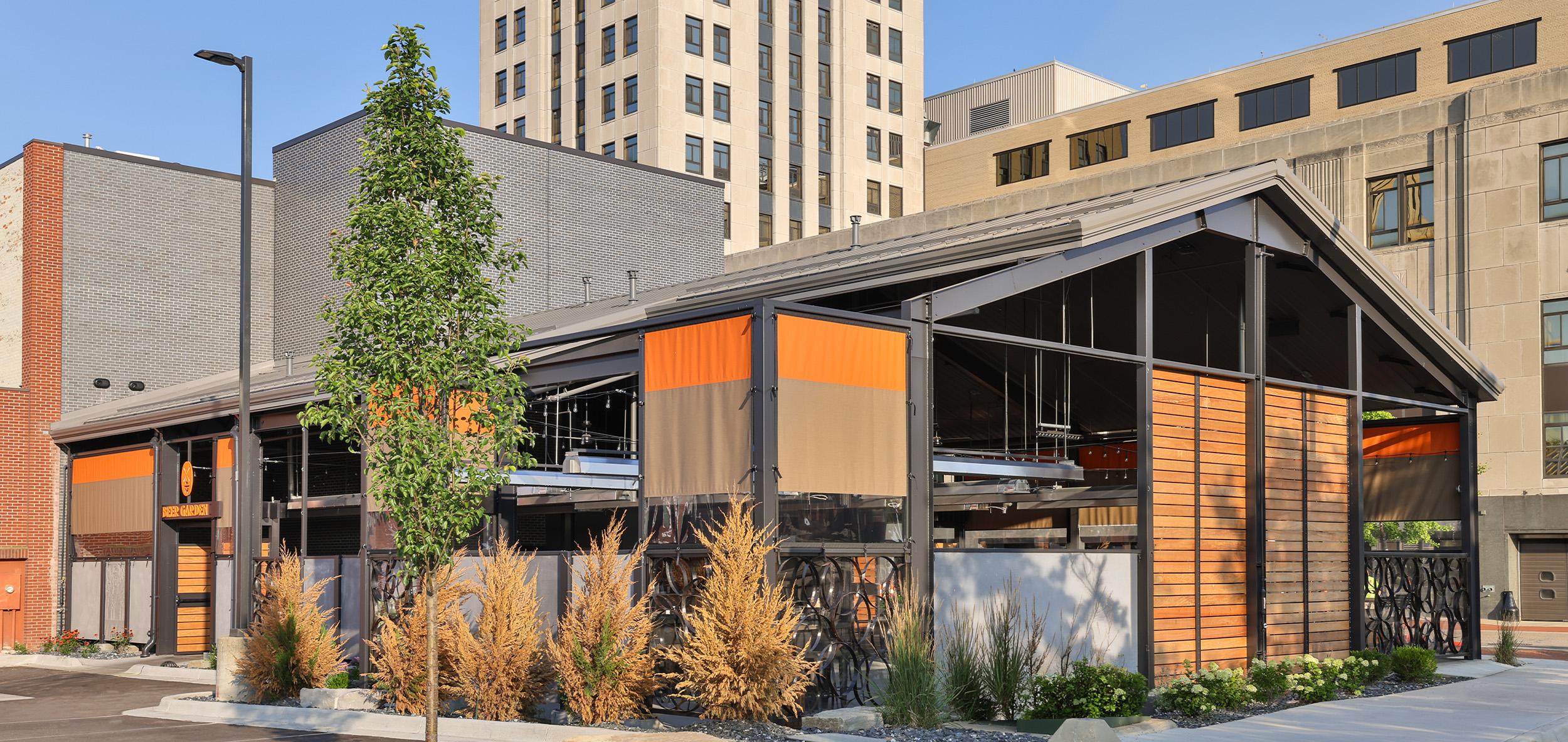 Contemporary urban pavilion with black steel framing, warm wood accents and open-air gathering spaces set among downtown buildings.