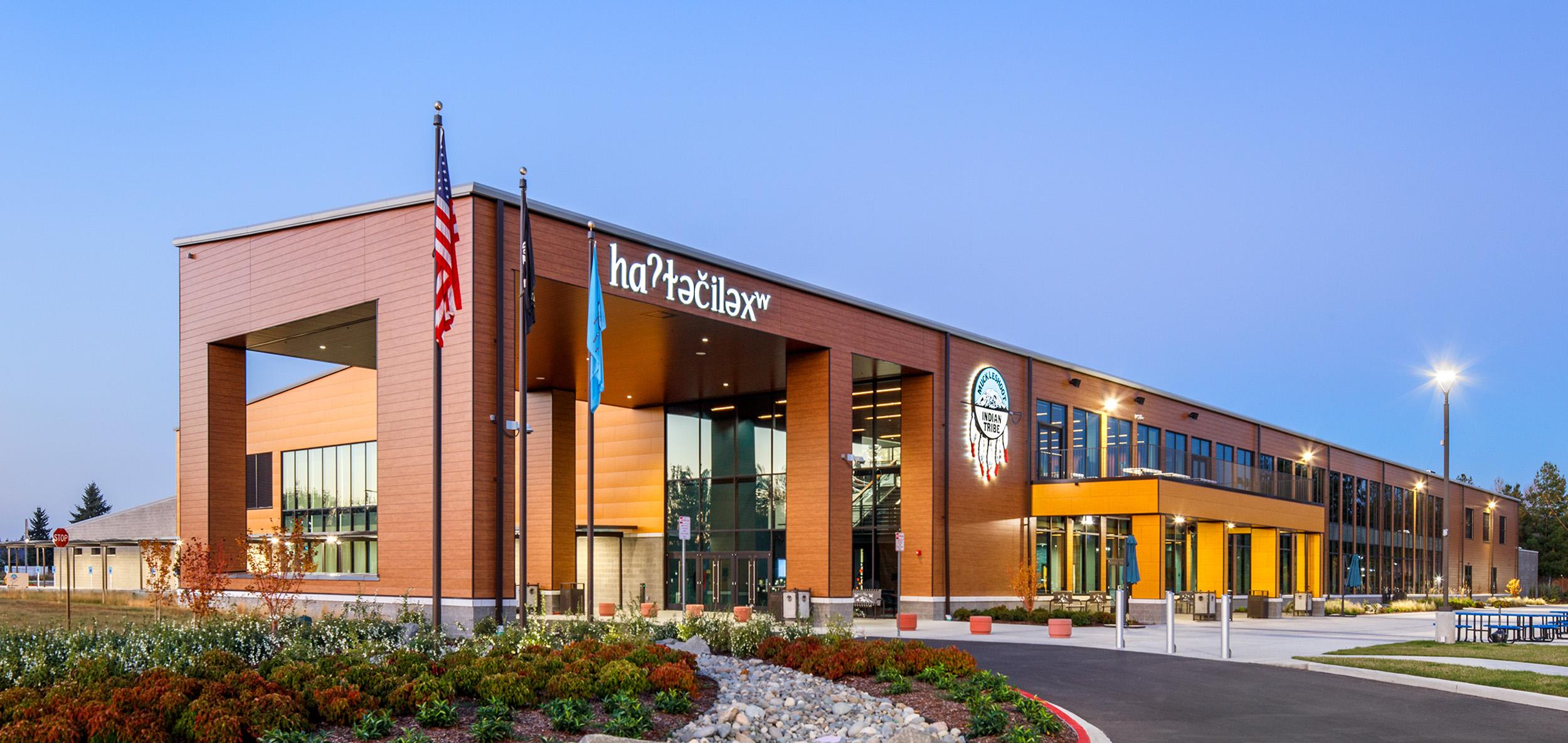 Contemporary civic building with warm wood-tone facade, expansive glass entry and landscaped frontage illuminated at dusk.