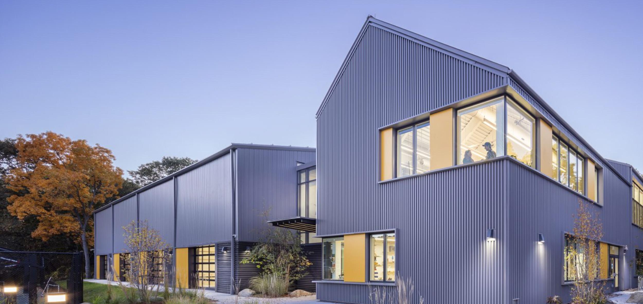 Contemporary dark gray education building with angular metal rooflines, warm interior lighting and landscaped walkways at dusk.