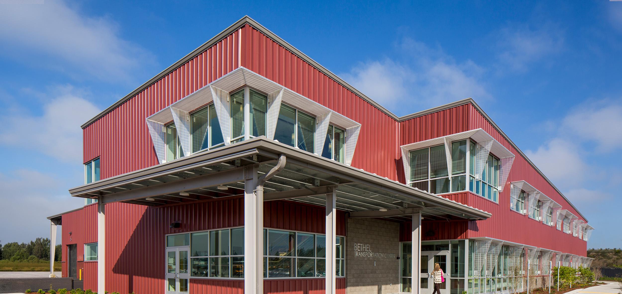 Contemporary red metal community building with dramatic cantilevered glass corners, covered entry walkway and bright blue sky backdrop.