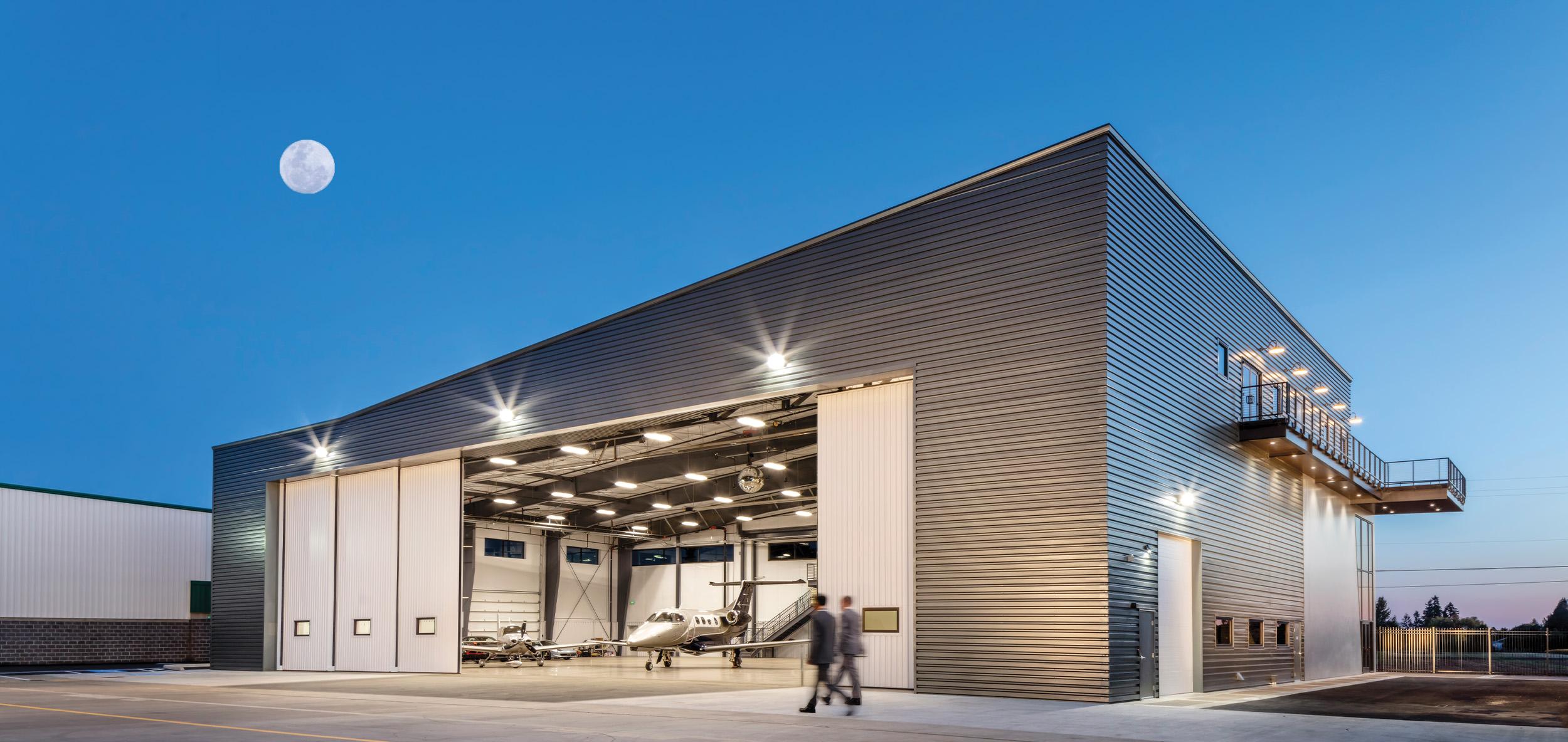 Modern aircraft hangar with expansive open service bays, illuminated interior workspace and sleek dark metal facade beneath a bright moonlit evening sky.