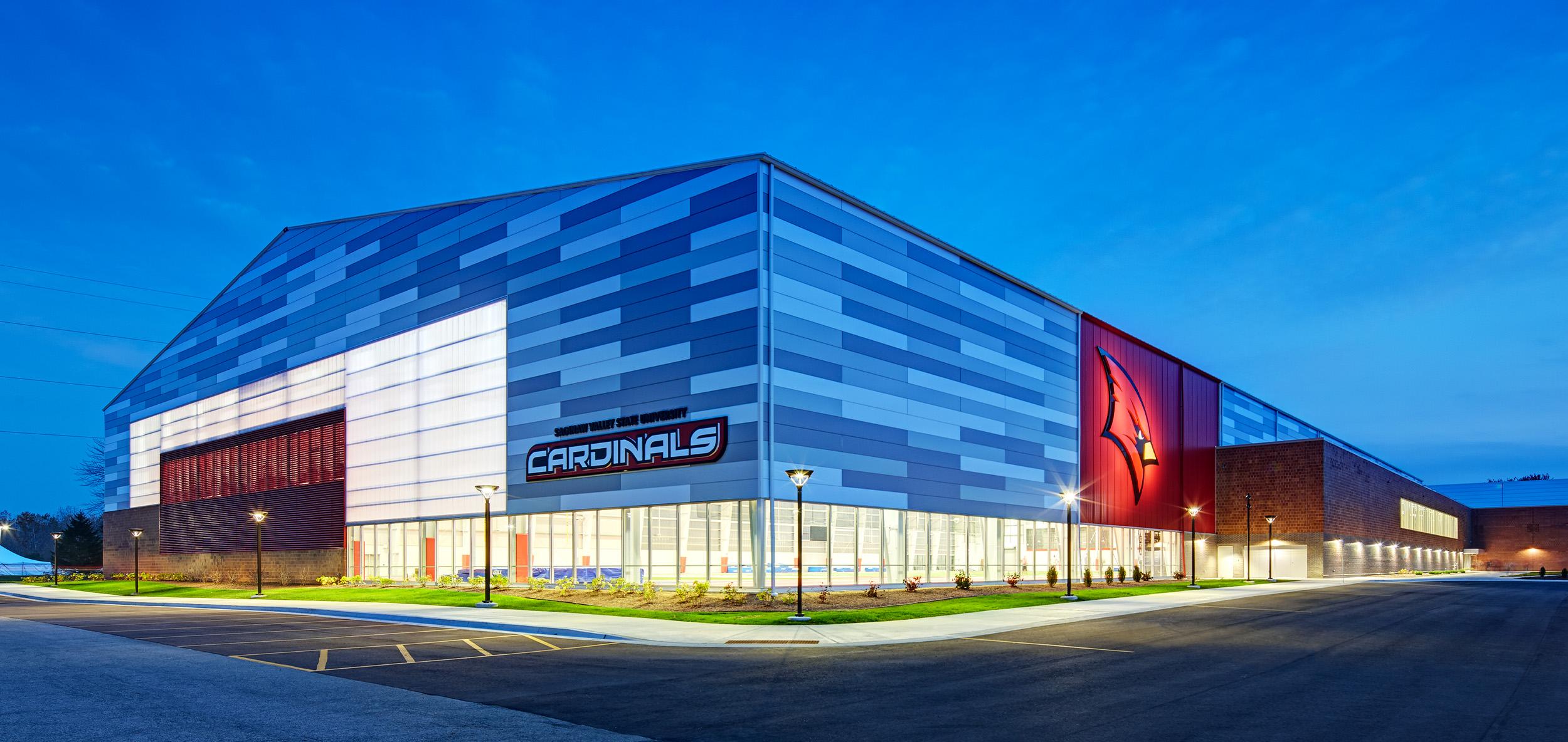 Large contemporary athletics facility with blue-gray metal panel facade, illuminated glass frontage and bold cardinal-themed entry accents at dusk.