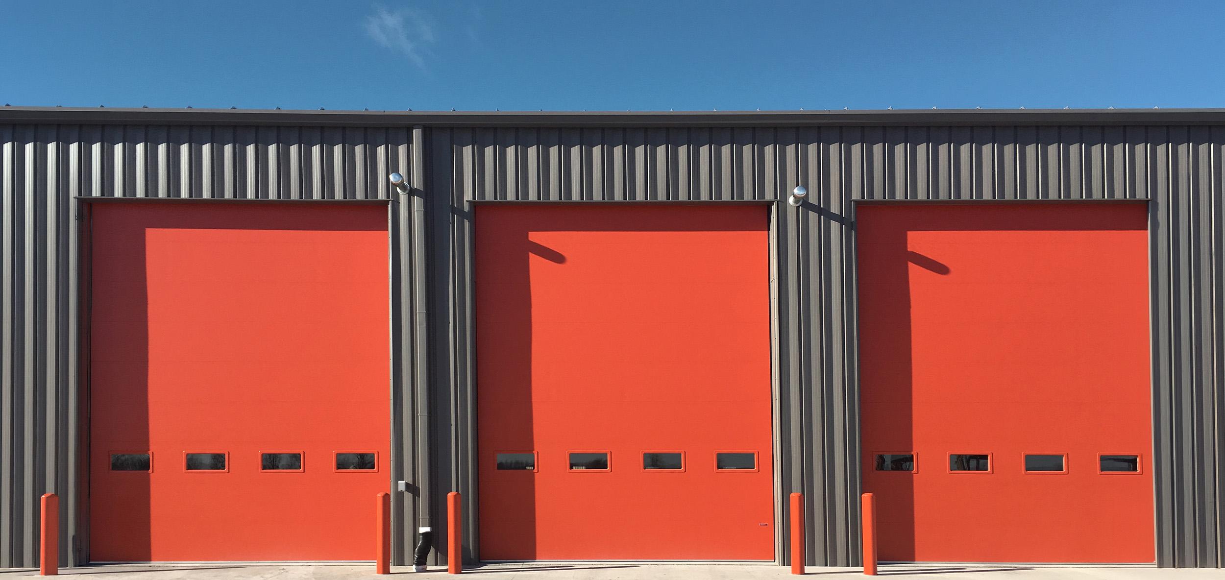 Industrial service facility with dark metal siding, bold red overhead doors and clean concrete apron beneath a clear blue sky.