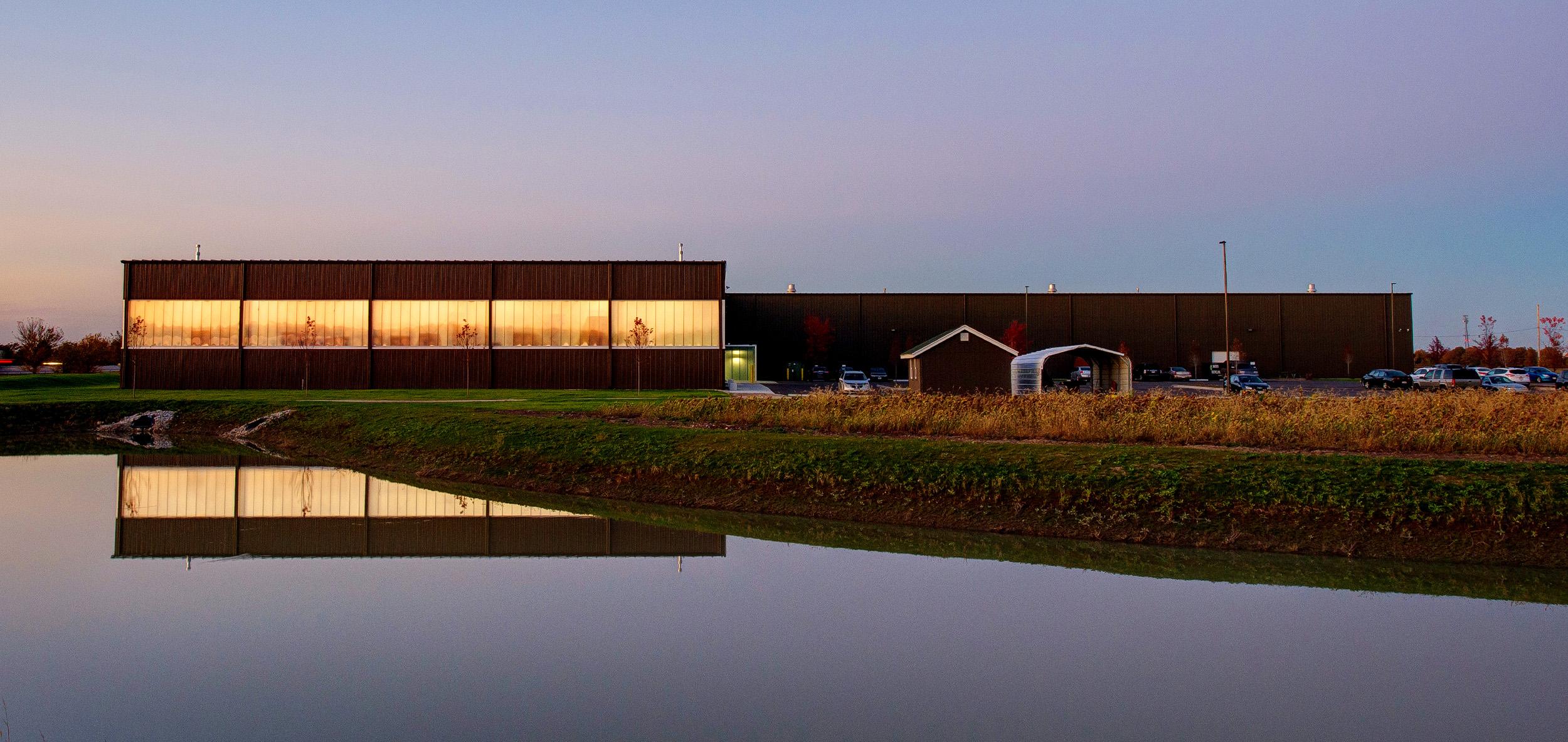 Long illuminated industrial building with reflective glass facade mirrored across a calm pond at dusk.