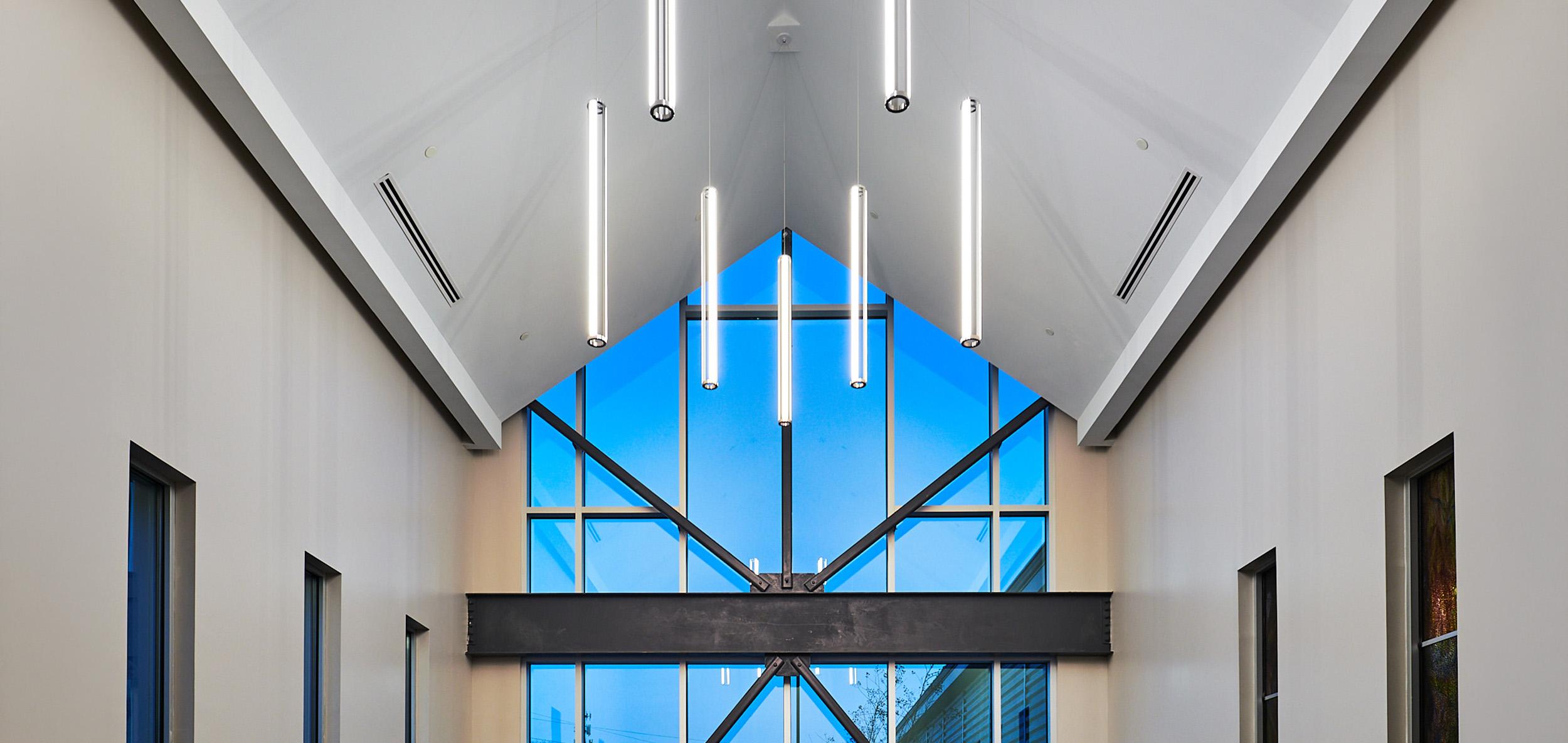 Modern chapel-style gathering space with vaulted ceiling, suspended linear lighting and expansive glass wall overlooking an urban skyline at dusk.