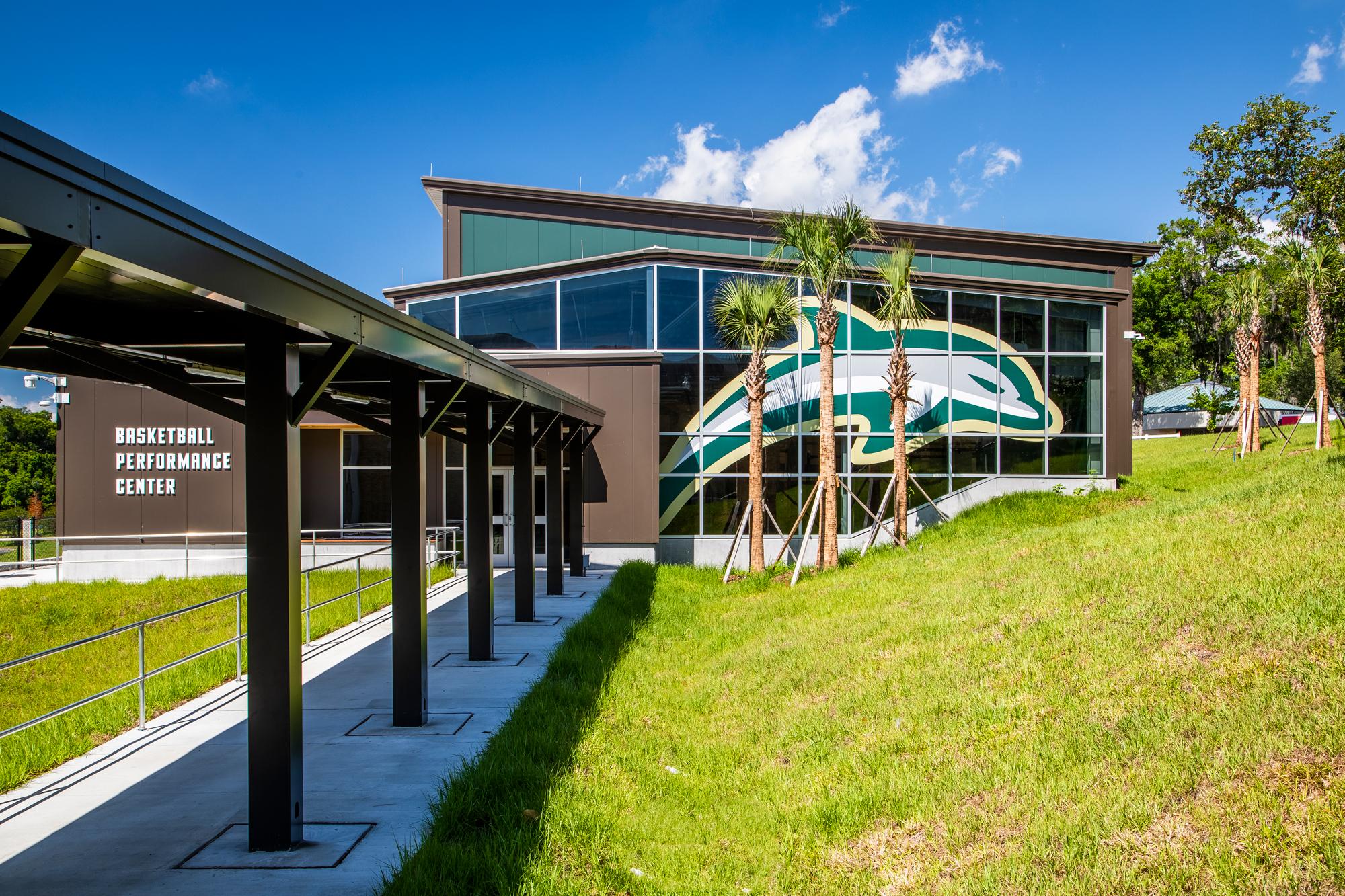 Green exterior of the Jacksonville University Basketball Performance Center featuring a large dolphin mascot graphic and covered walkway entrance.