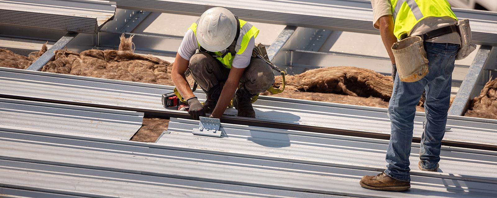 Construction professionals installing a standing seam metal roof system, demonstrating active field coordination and precision roof panel placement during construction.