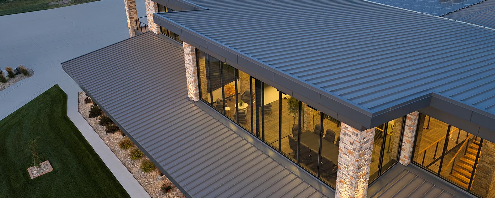 Aerial view of a modern commercial building featuring a gray standing seam roof, expansive glass walls and stone-accented columns showcasing integrated roof and facade design.