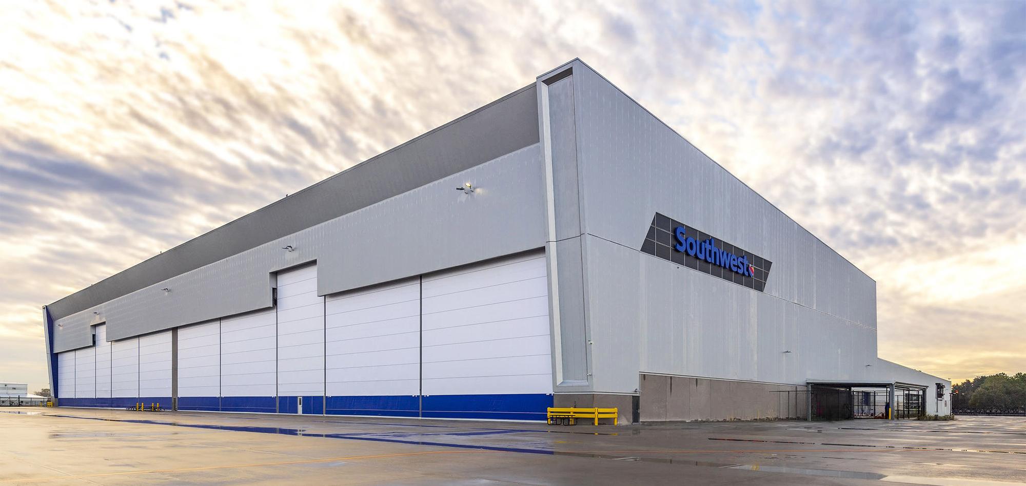 Large white and gray industrial building with multiple tall overhead doors, blue trim and a Sundt sign beneath a partly cloudy sky.