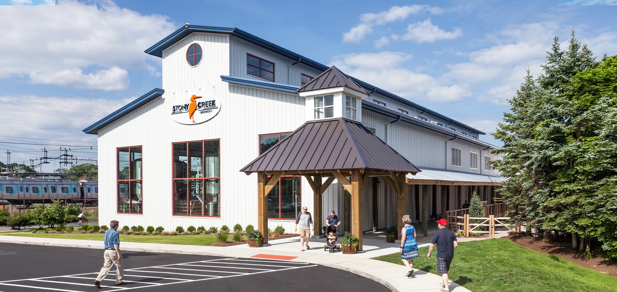 Contemporary community recreation center with white facade, metal rooflines, covered entry pavilion and visitors gathering along landscaped walkways beneath a bright blue sky.