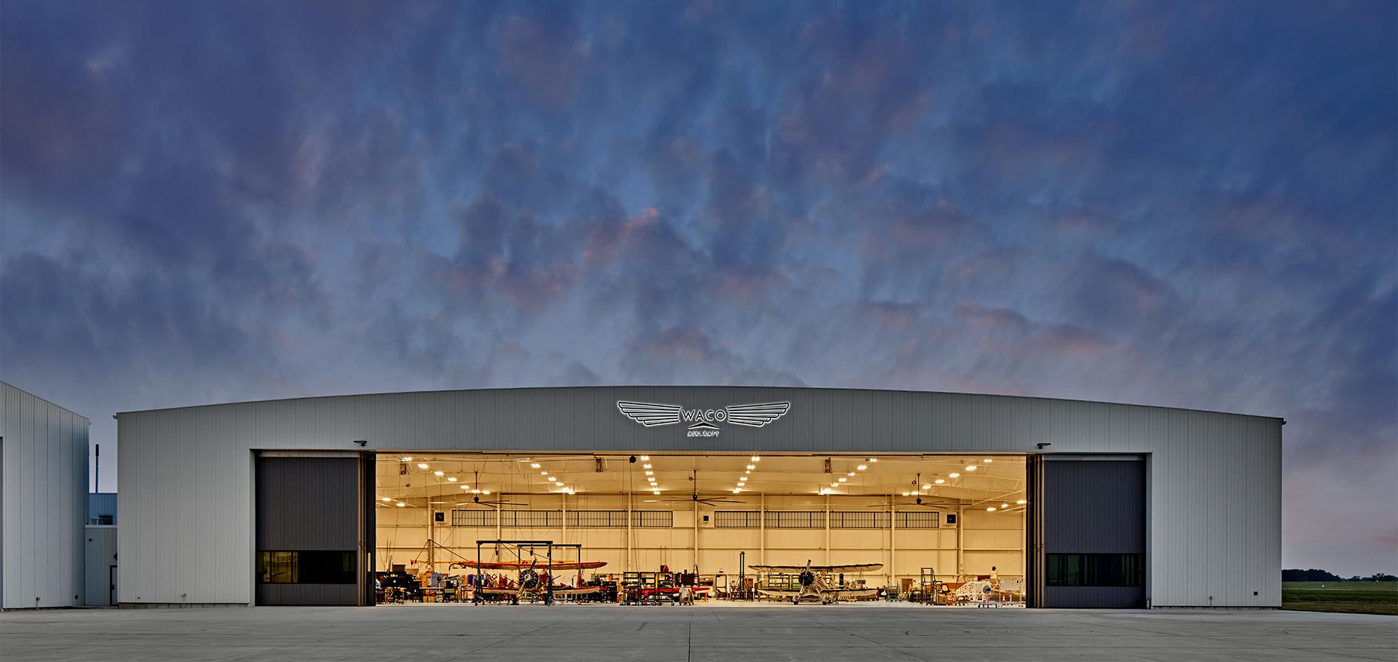 Front exterior view of the WACO Aircraft Corp. facility featuring a large open aircraft hangar and integrated aviation production environment designed to support coordinated manufacturing, assembly and customer engagement operations.
