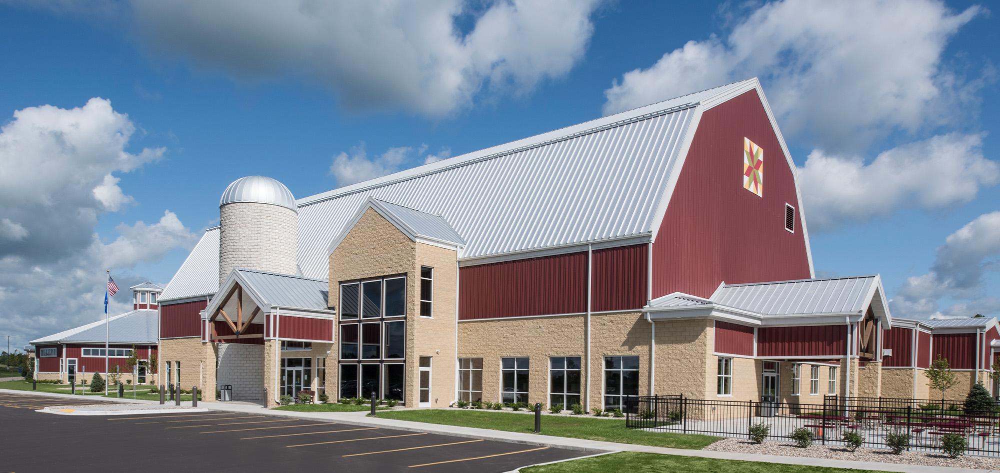 Red and tan agricultural facility with a white metal roof, central silo feature and expansive glass entry designed to serve agricultural operations and community gathering spaces beneath a bright sky with scattered clouds.