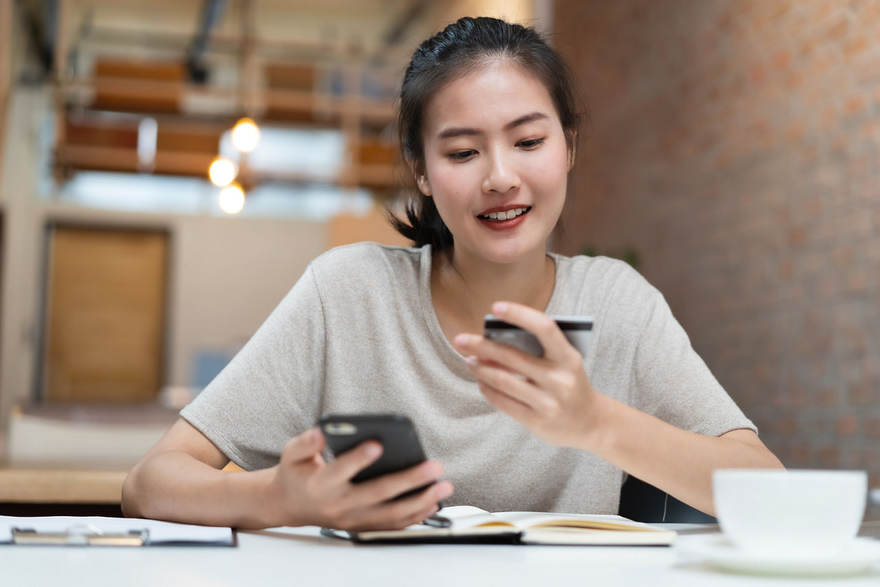 Happy young asian woman buying stuff from online retail and paying bills via banking application. Girl looking at credit card in her hand and fill data to mobile phone to complete purchese order..