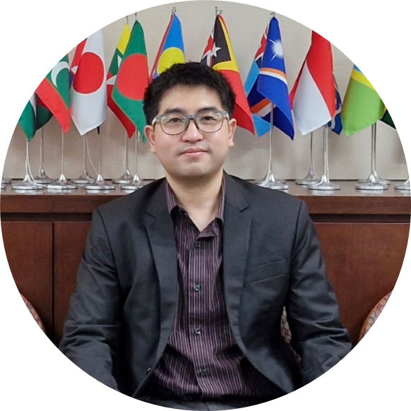 ID photo of Naruedom Sinudom a young man with black hair and black eyes and light complexion wearing a grey suit jacket and scarlet shirt with multiple country flags in the background