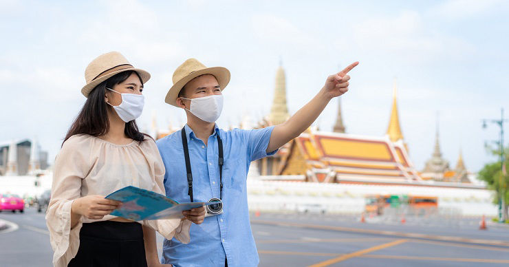 Asian couple happy tourists to travel wearing mask to protect from Covid-19 on they holidays and holding travel map and pointing in Wat Phra Kaew Temple in Bangkok, Thailand