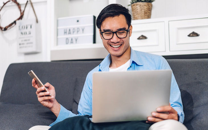 Young smiling asian man relaxing using laptop computer working and video conference meeting at home.Young creative man looking at screen typing message with smartphone.work from home concept