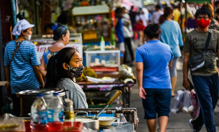 BANGKOK, THAILAND - MAY, 2020: thai female street vendor wearing a mask, looking around and waiting for customers in a busy crowded market street on Ramkamhaeng Rd.