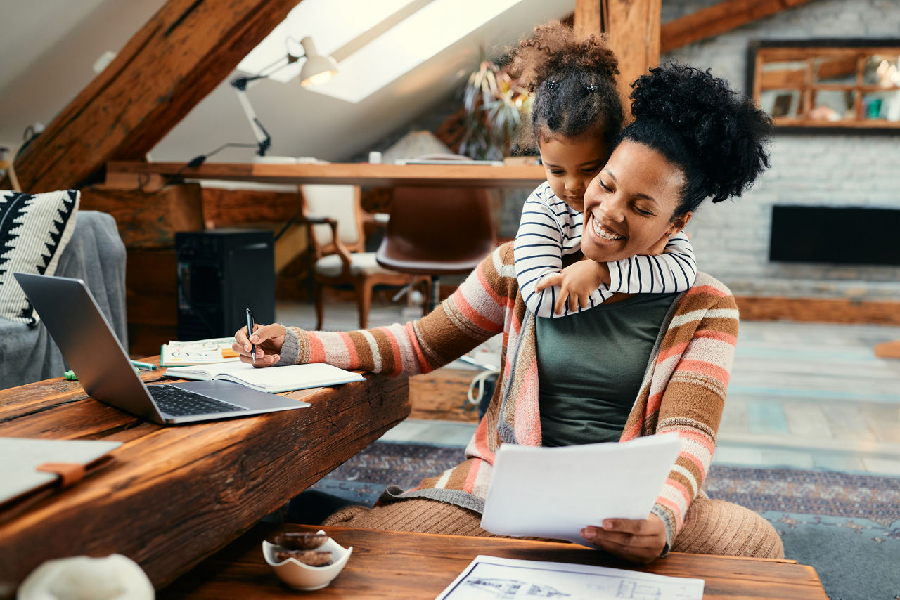 Happy African American mother using laptop and working on paperwork while her daughter is embracing her at home.