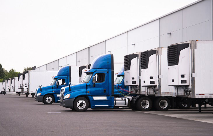 A day cab big rigs semi trucks with a reefer trailers stand near the gate of the warehouse next to other reefer trailers that are loaded and unloaded to deliver perishable and frozen food to consumers