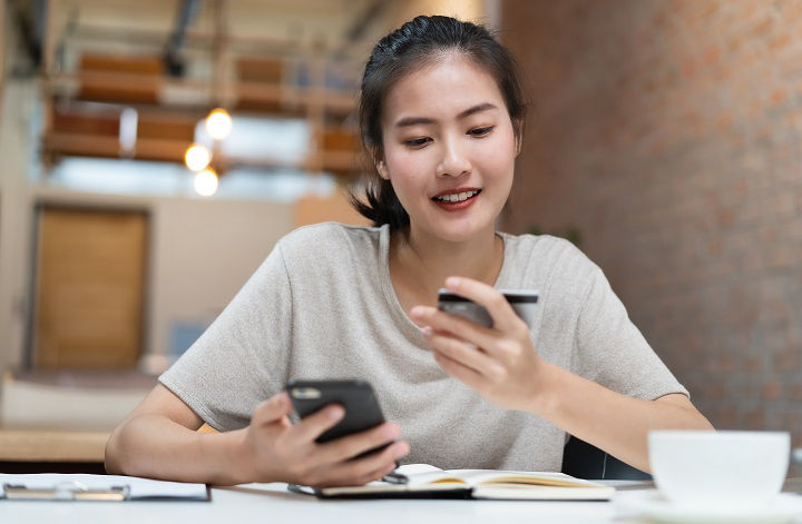 Happy young asian woman buying stuff from online retail and paying bills via banking application. Girl looking at credit card in her hand and fill data to mobile phone to complete purchese order..