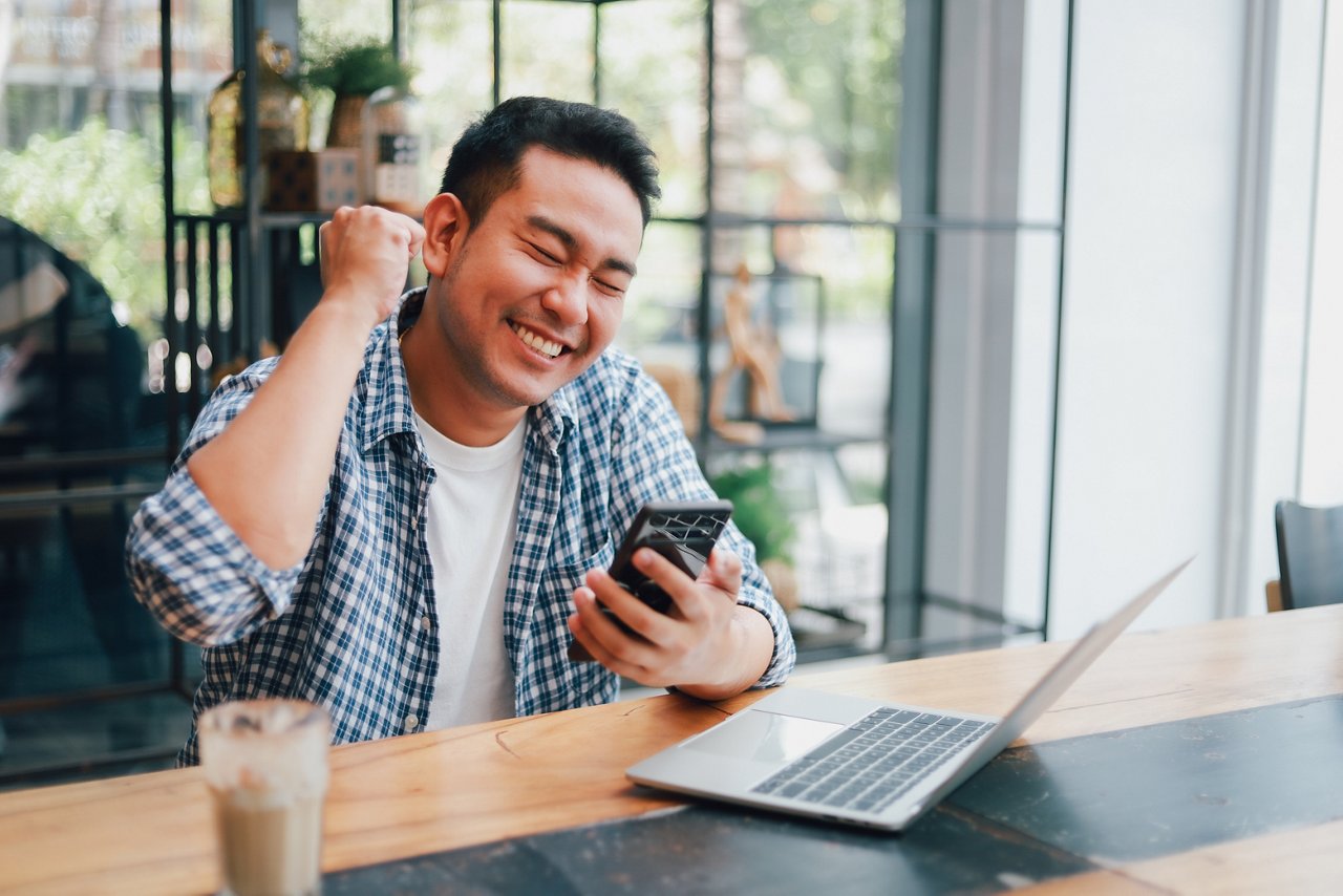 Asian young man in blue shirt working with laptop computer and mobile phone with online job or shopping online happy and smile face in coffee shop cafe