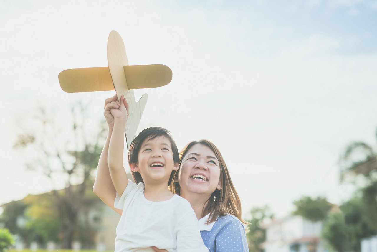 Cute Asian mother and son playing cardboard airplane together in the park outdoors
