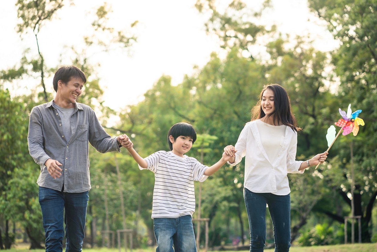 Asian happy young family walk in park in nice weather. Parent hold son hand and smile. Mother hold colorful wind turbine. People love to do relax activity together. Family slow life lifestyle concept
