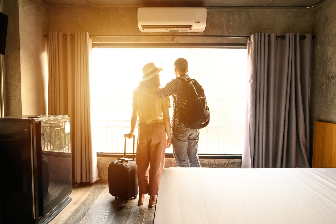 Portrait of young couple tourist standing nearly window, looking to beautiful view outside in hotel/resort bedroom after check-in. Conceptual of couple travel and vacation in their honeymoon period.