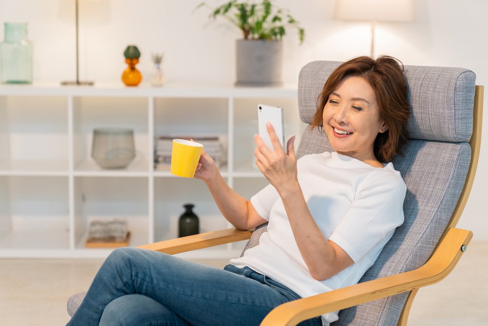 A woman looking at her phone smiling while also holding a cup of coffee in a living area.