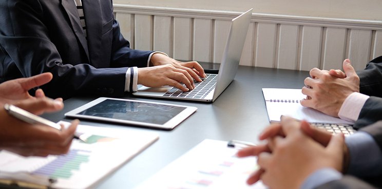 Hands of people in a meeting with laptop, tablet, notebook, and papers on a table.