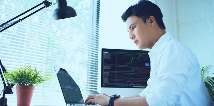 A man typing on his laptop with a desktop monitor showing graphs.