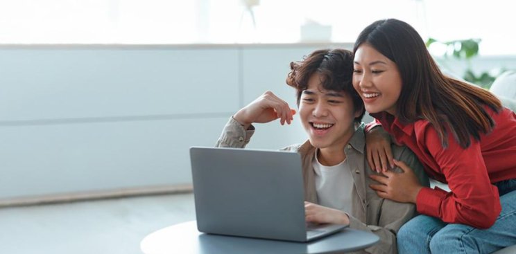 A young man and woman smiling as they look at a device's screen inside a well-lit living area.