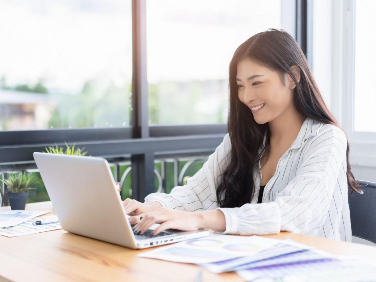 A woman smiling at her laptop near a glass window.